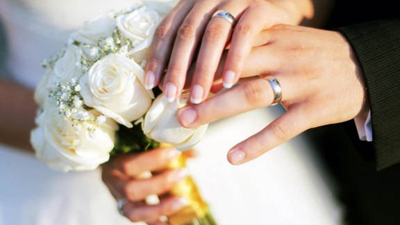 Nahaufnahme zweier Hände mit Eheringen, die auf einem Brautstrauß aus weißen Rosen ruhen – symbolisch für Eheschließung und Hochzeitsfeier.
